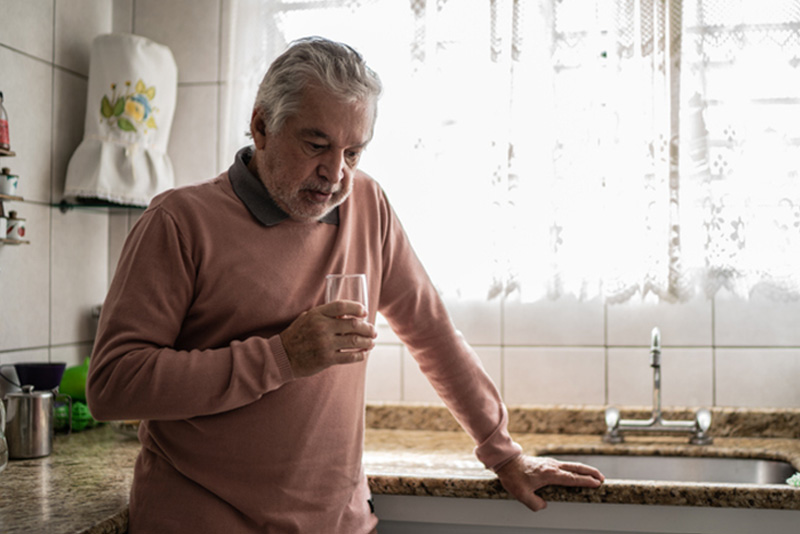 A senior man suffering from depression and cardiovascular disease stands in the kitchen holding a glass of water.